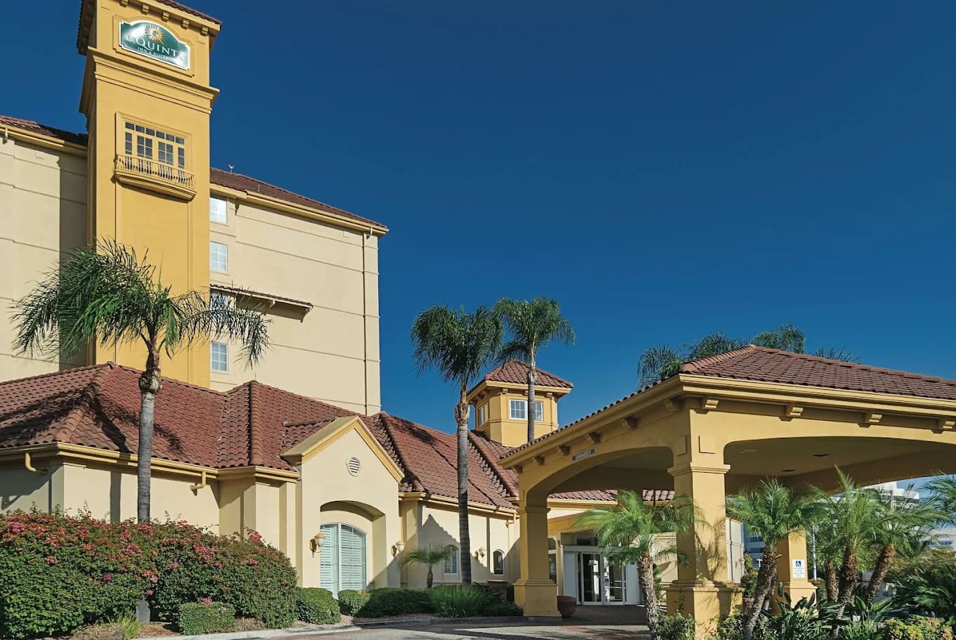 Yellow stucco La Quinta Inn & Suites exterior with red tile roofs, a covered entrance and palm trees under a blue sky