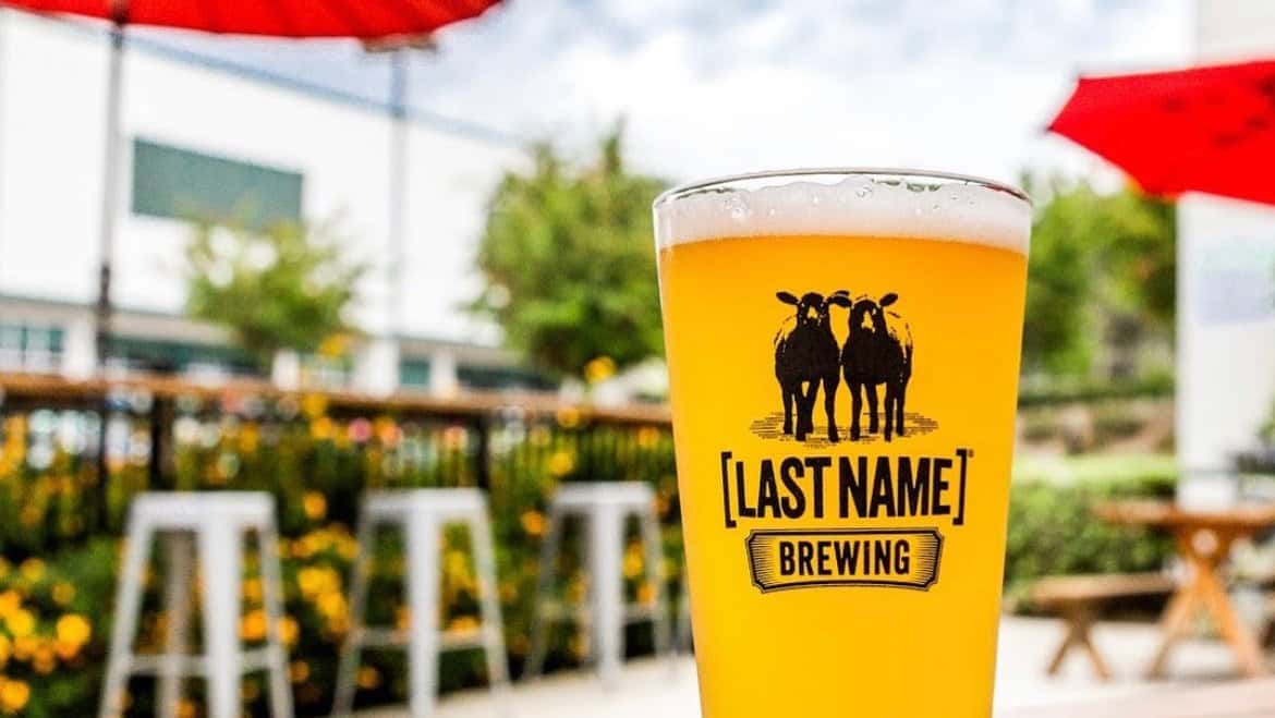 Pint glass with the Last Name Brewing logo held up in front of an outdoor patio with red umbrellas
