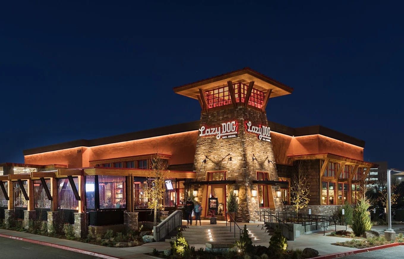 The Lazy Dog exterior at dusk, a stone-and-timber lodge front lit warmly under a deep blue sky