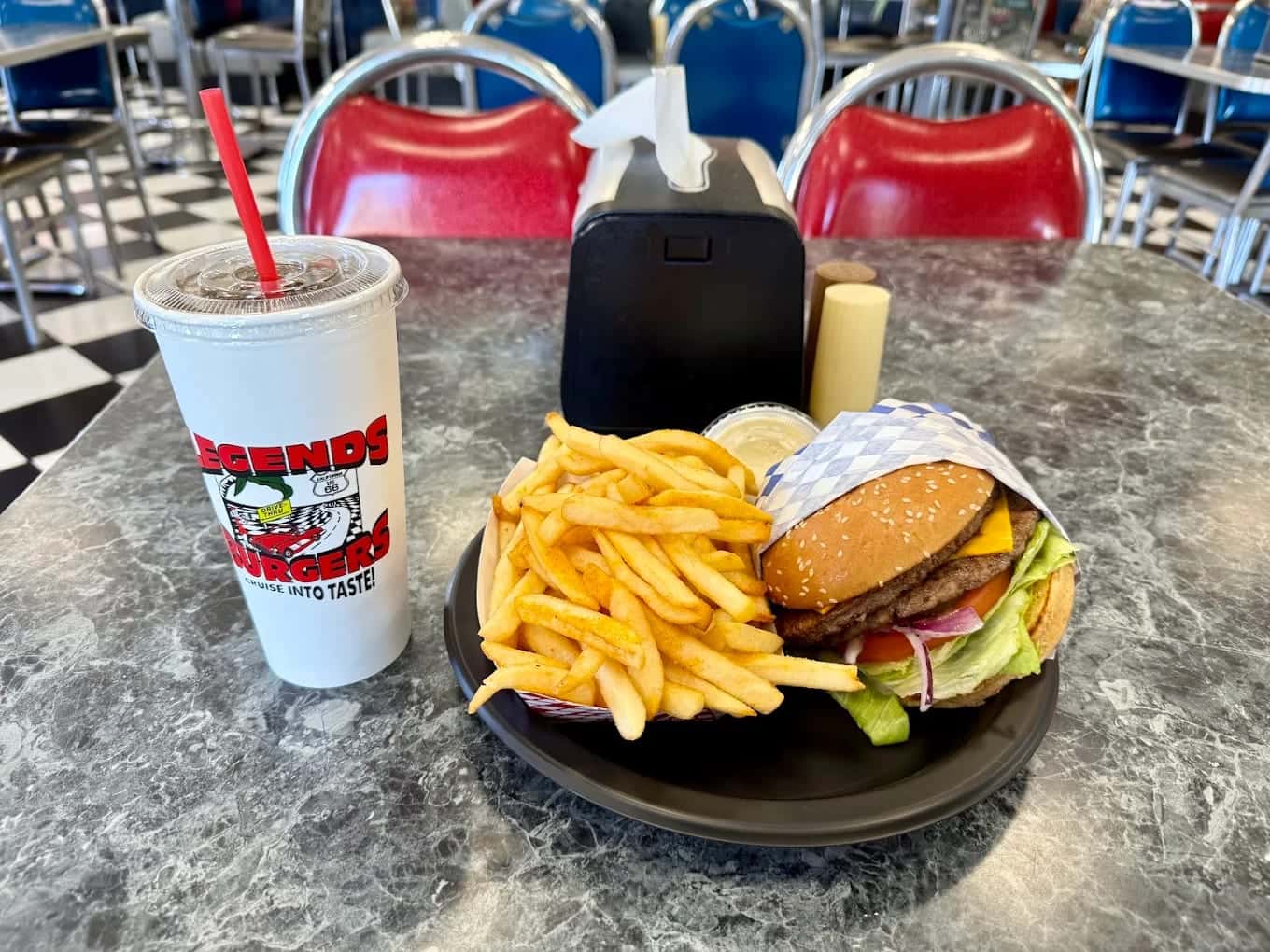 A cheeseburger and fries on a diner table beside a Legends Burgers cup, red booths behind