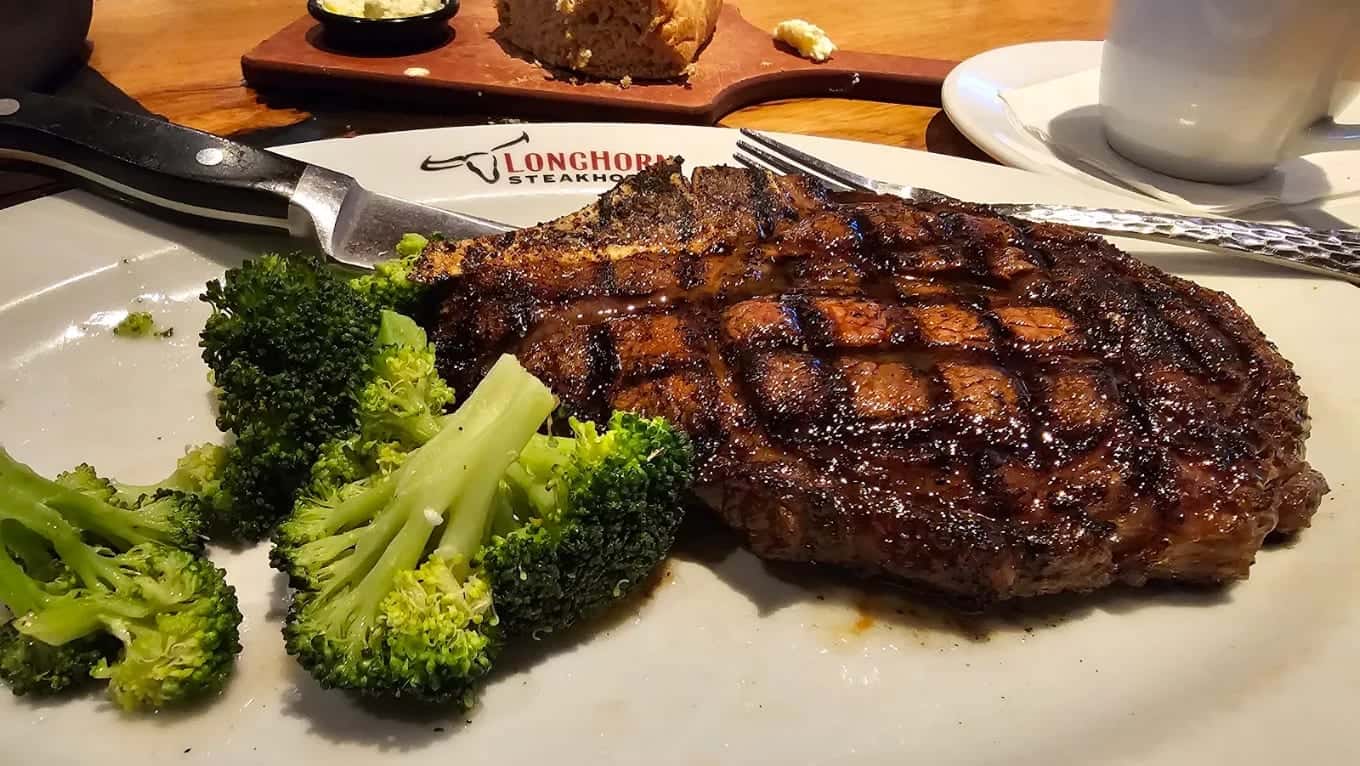 A grill-marked steak with broccoli on a white plate, a steak knife and a bread board behind it