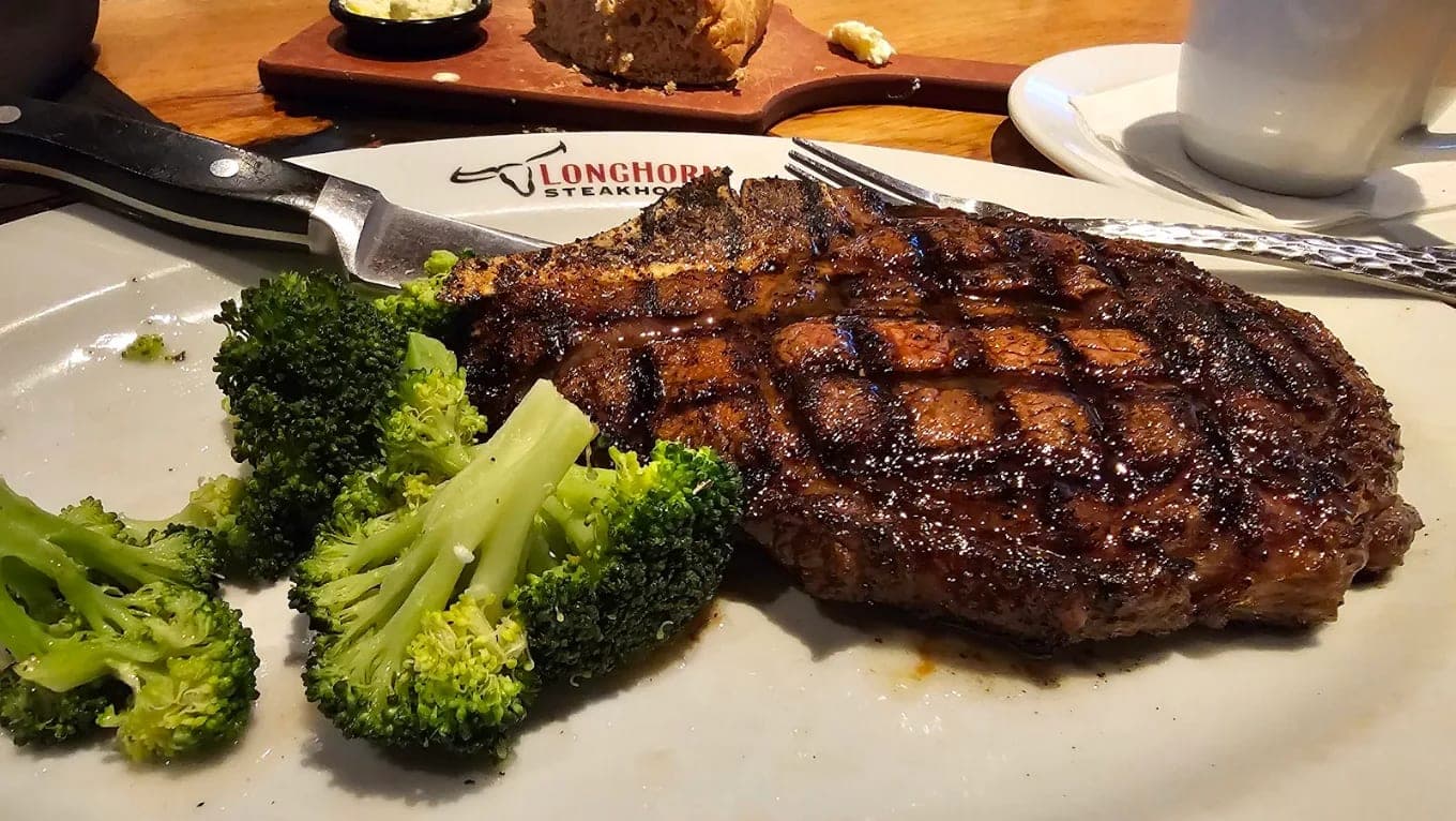 A grill-marked steak with broccoli on a white plate, a steak knife and a bread board behind it