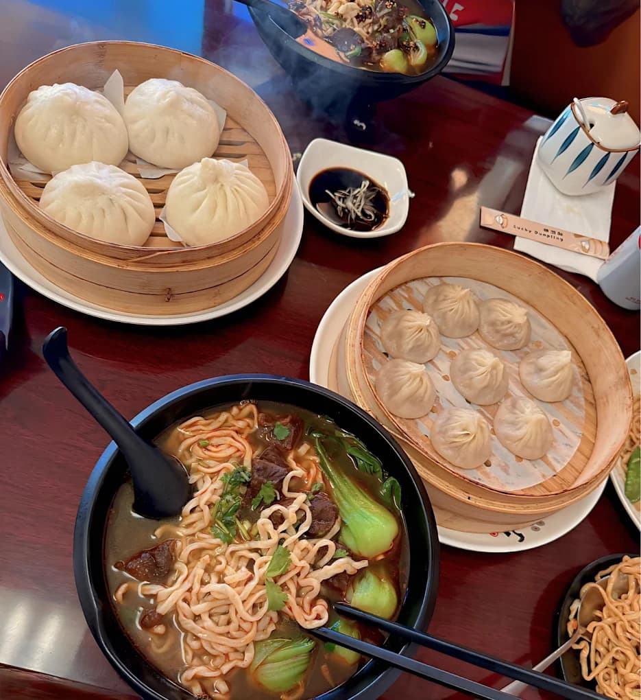 Steamed buns and dumplings in bamboo baskets beside a bowl of beef noodle soup at Lucky Dumpling