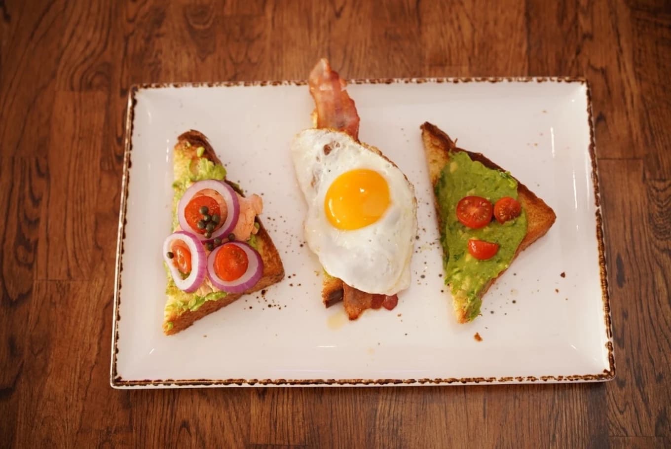 Three toast triangles topped with avocado, a fried egg and bacon on a white plate at Madres Brunch
