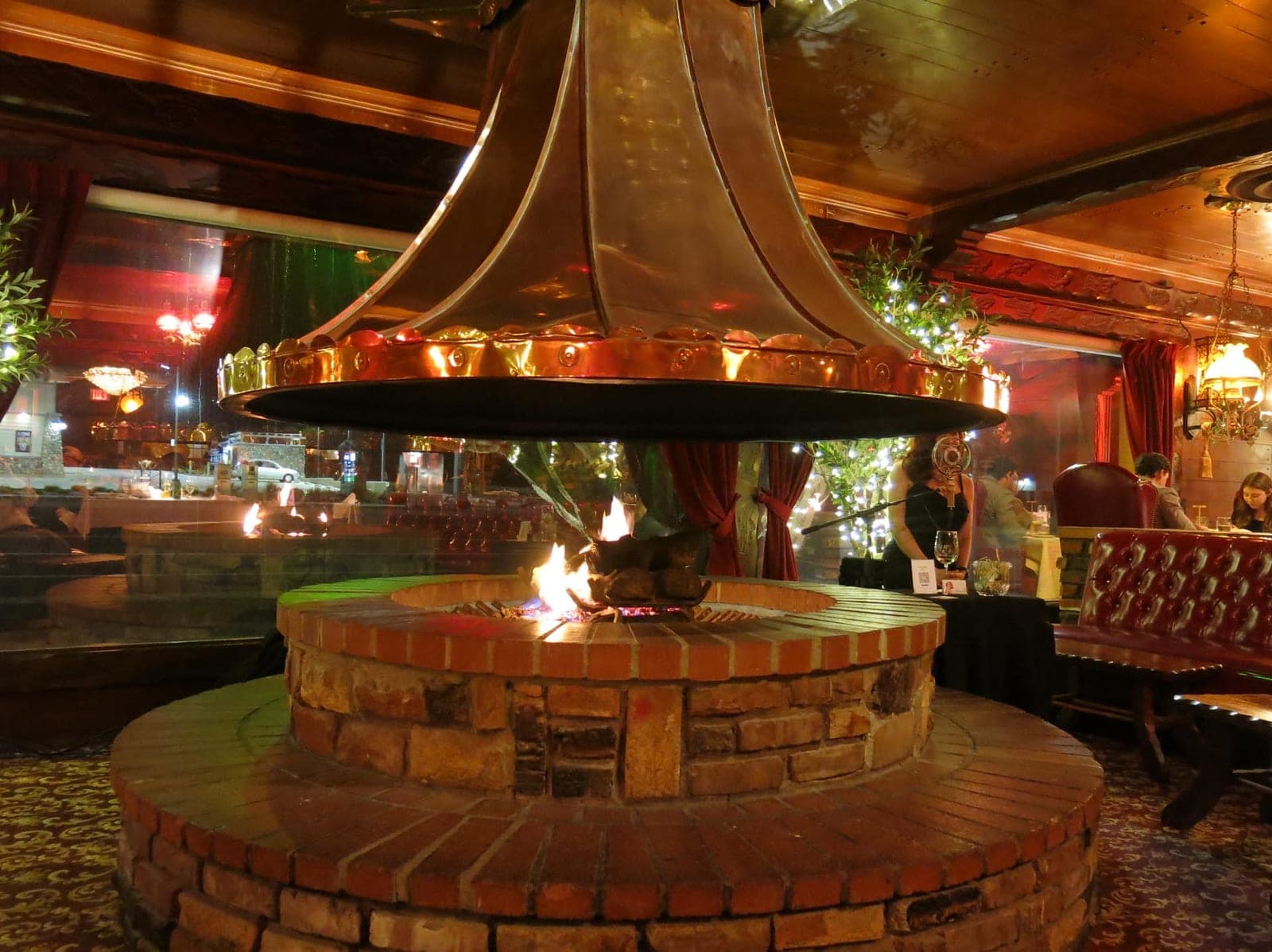 The circular brick fire pit in the Magic Lamp Inn dining room, burning under a polished copper hood with red leather booths beyond