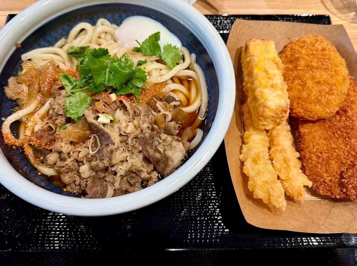 Beef udon bowl with a soft egg and cilantro beside tempura and croquettes on a tray at Marugame Udon
