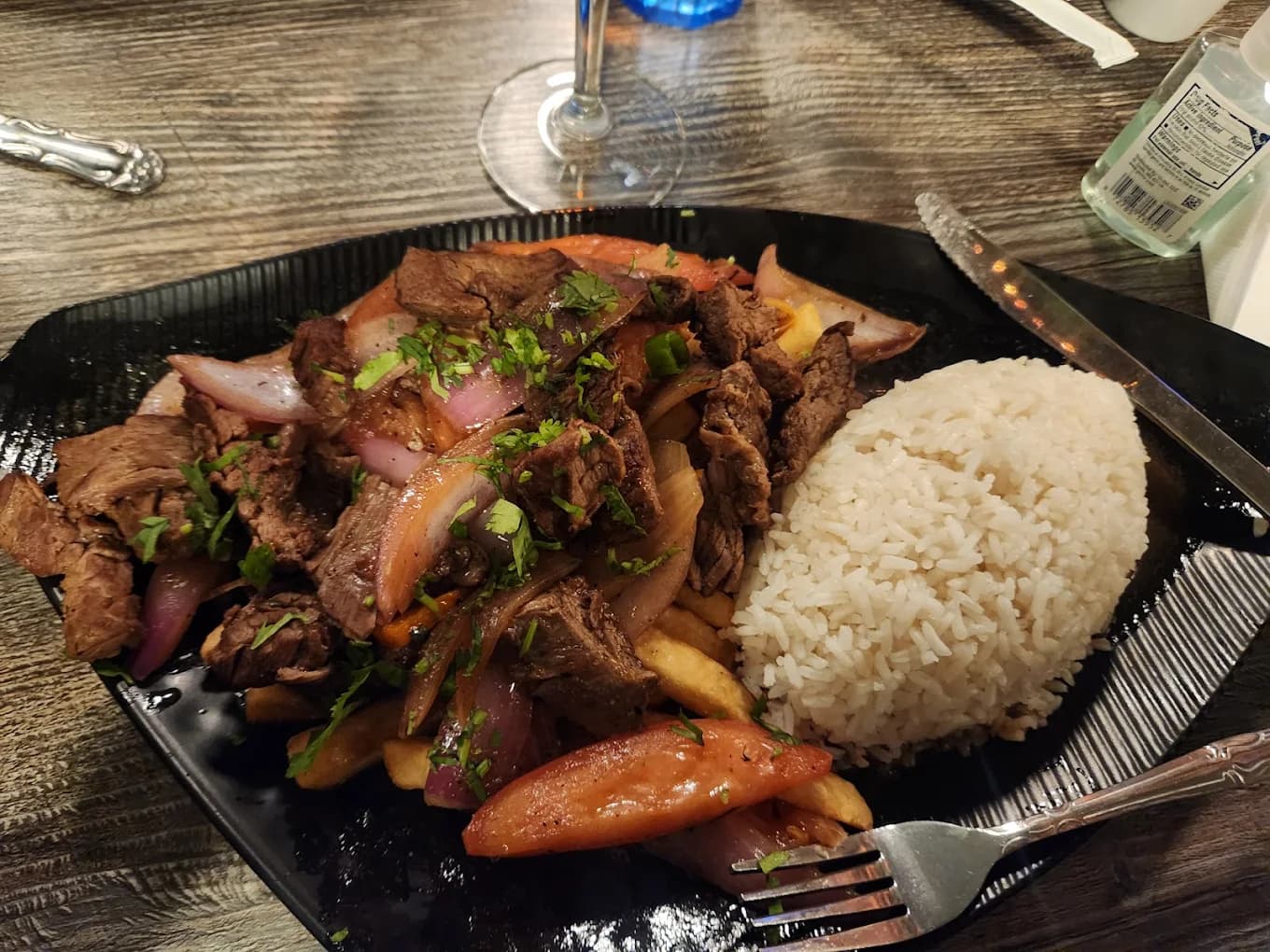 Sliced beef stir-fried with red onion, tomato and cilantro beside a mound of white rice on a black plate at Micas