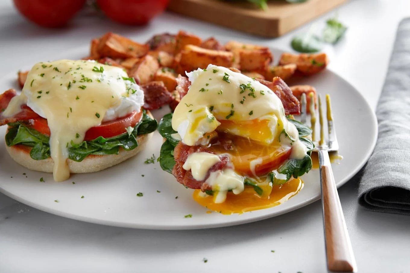 Two eggs Benedict on English muffins with spinach, tomato and bacon under hollandaise, one yolk broken and running