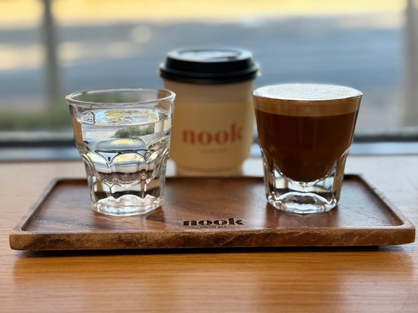 Espresso drink, glass of water and a branded paper cup on a wooden tray at Nook Coffee Bar