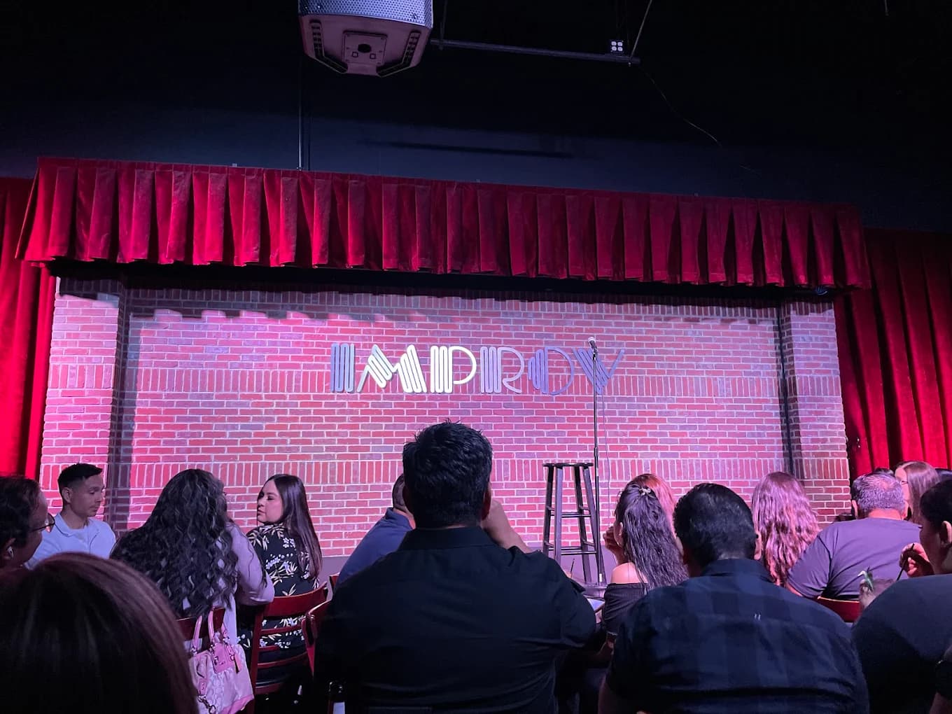 Brick stage with Improv sign, microphone and stool under red curtains at Ontario Improv, audience seated
