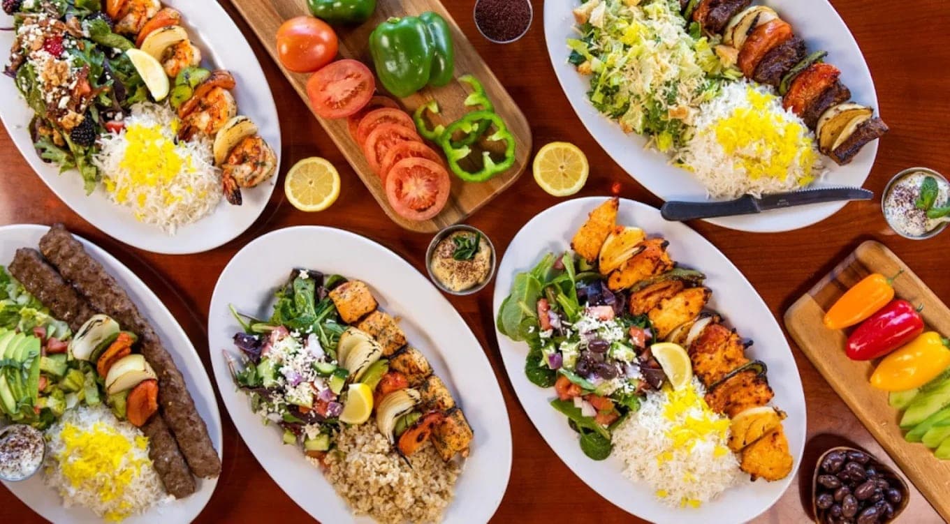 An overhead spread of kabob plates with saffron rice and salads, peppers and tomatoes on boards between them