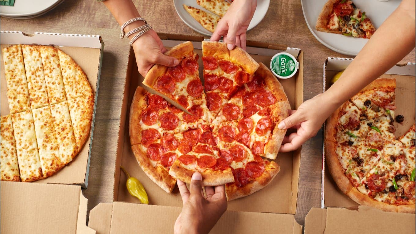 Hands reaching for slices of pepperoni pizza in open boxes at Papa Johns Pizza, cheese sticks alongside