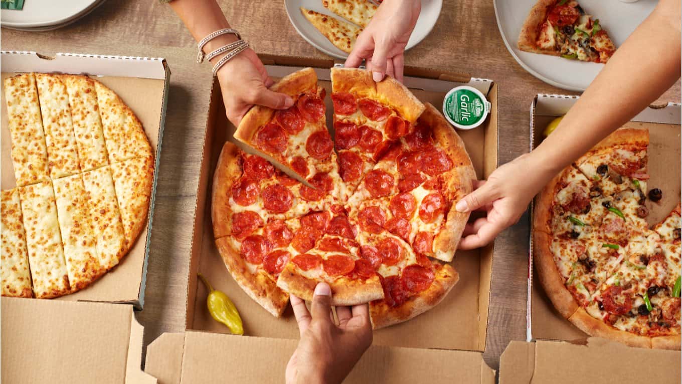 Hands reaching for slices of pepperoni pizza in open boxes at Papa Johns Pizza, cheese sticks alongside