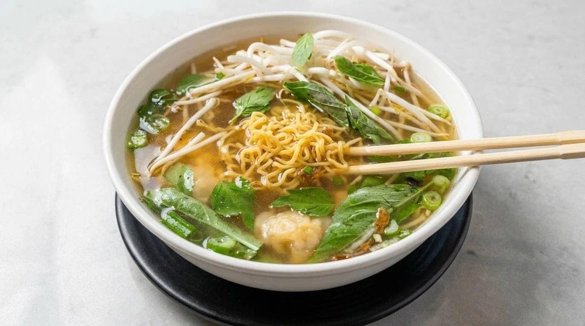 A bowl of pho with egg noodles, wontons, bean sprouts, herbs and spring onion, chopsticks resting across the rim