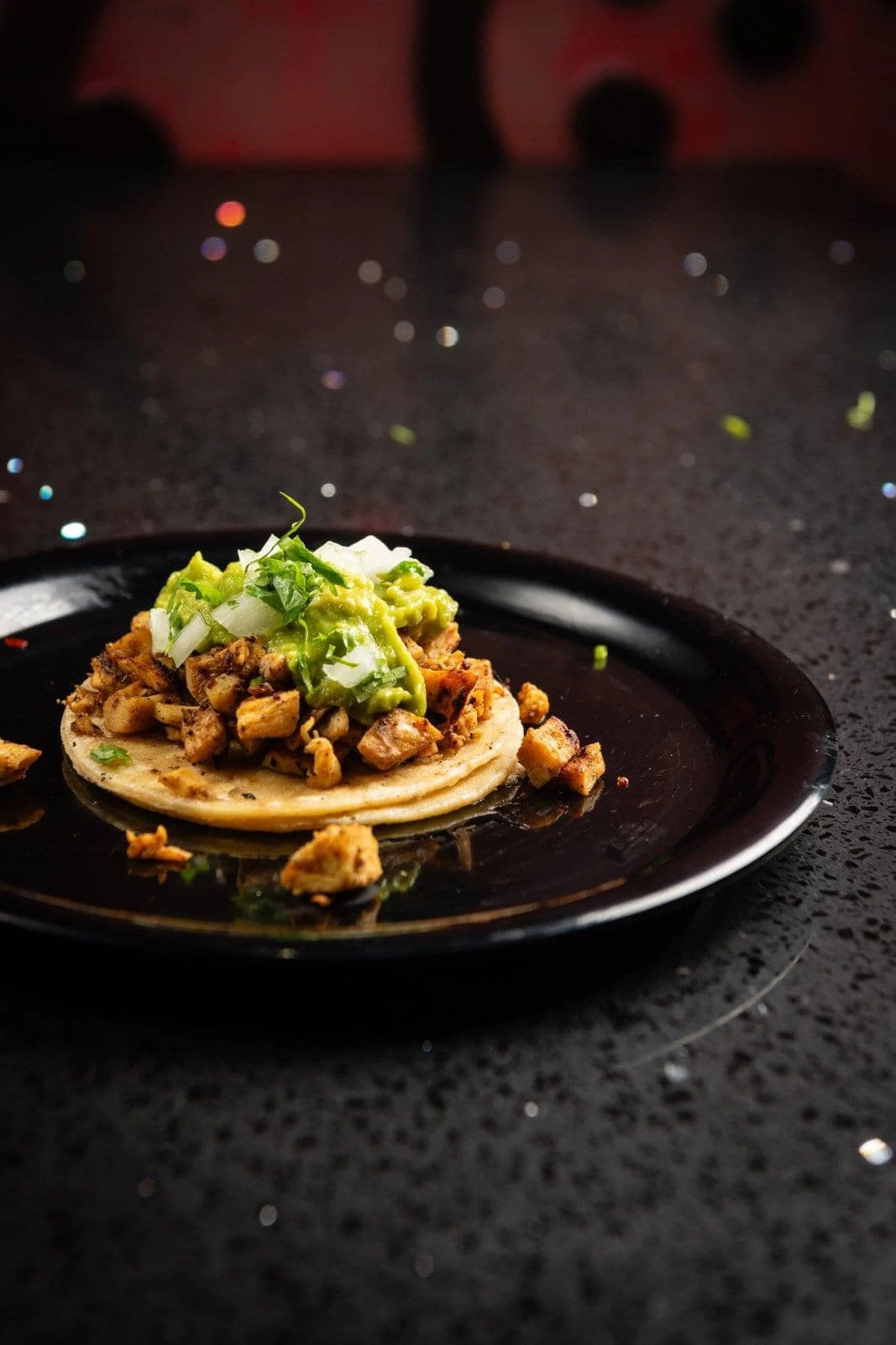 A taco on a double corn tortilla, chopped meat under guacamole, onion and cilantro on a dark plate