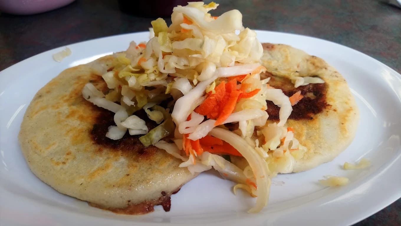 Two pupusas topped with curtido cabbage slaw on a white plate at Pupusería Doña María