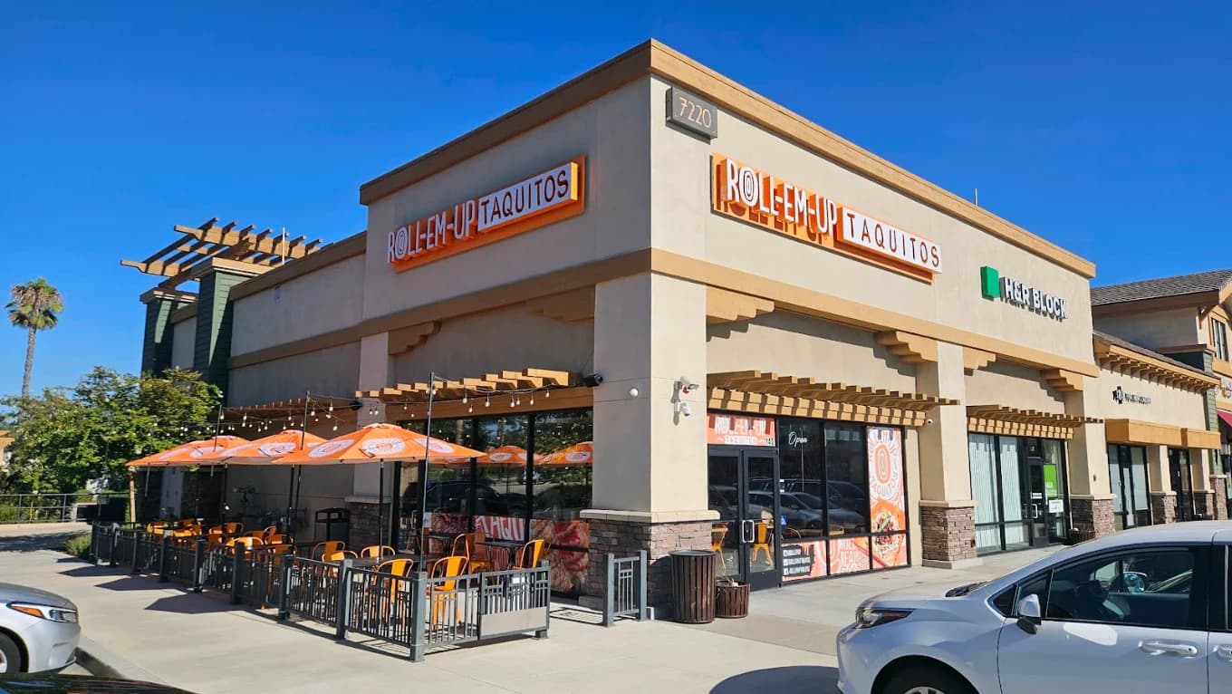 Storefront of Roll Em Up Taquitos with orange signs and a fenced patio with umbrellas