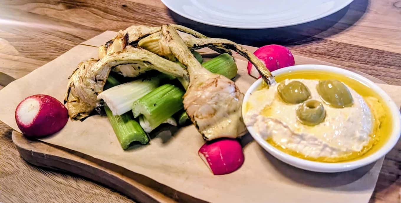 Grilled artichokes, celery and radishes on a board with a bowl of hummus and olives at Seasons 52