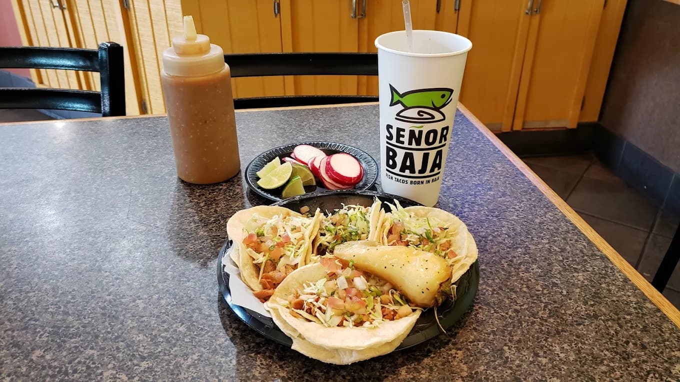 Three tacos on a tray with radishes, lime, a squeeze bottle of sauce and a branded cup at Senor Baja