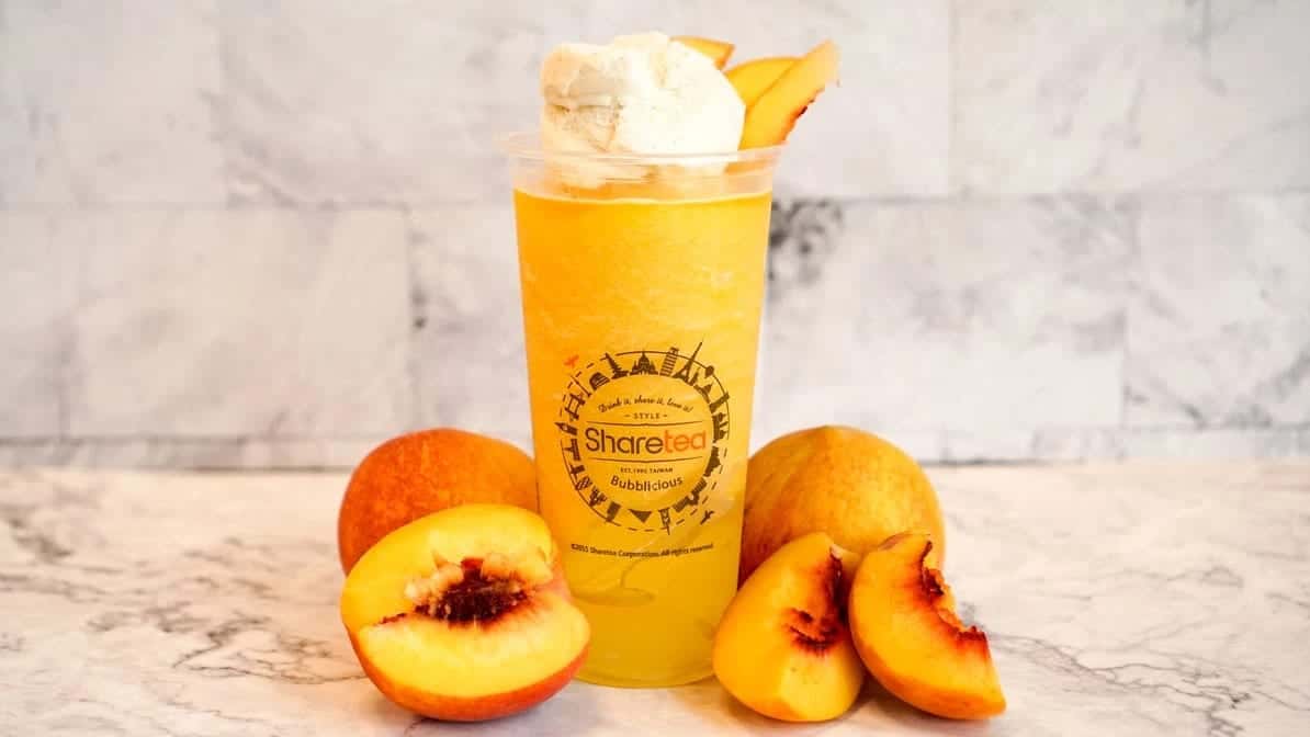 A cup of peach slush topped with cream, cut peaches arranged around its base on a marble counter