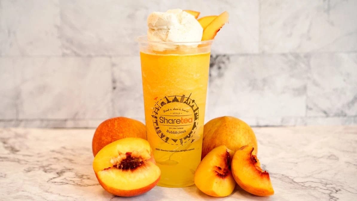 A cup of peach slush topped with cream, cut peaches arranged around its base on a marble counter