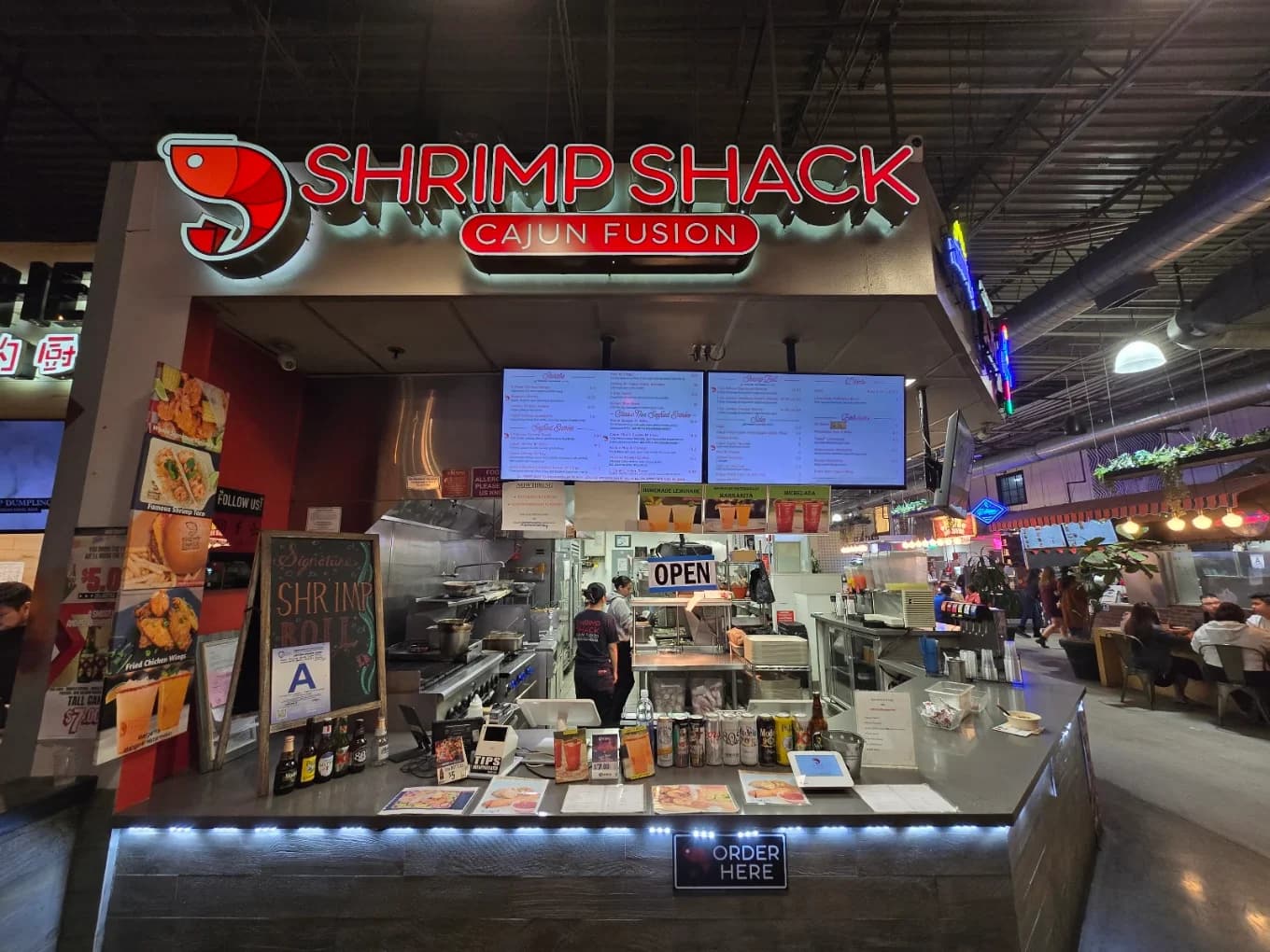 Food hall counter with lit red sign, digital menu boards and chalkboard at Shrimp Shack Cajun Fusion