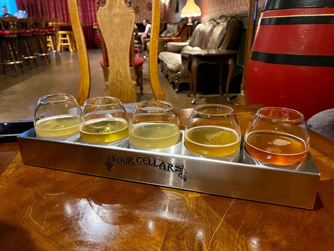 Flight of five beers in a branded metal tray with a red barrel and armchairs behind at Sour Cellars
