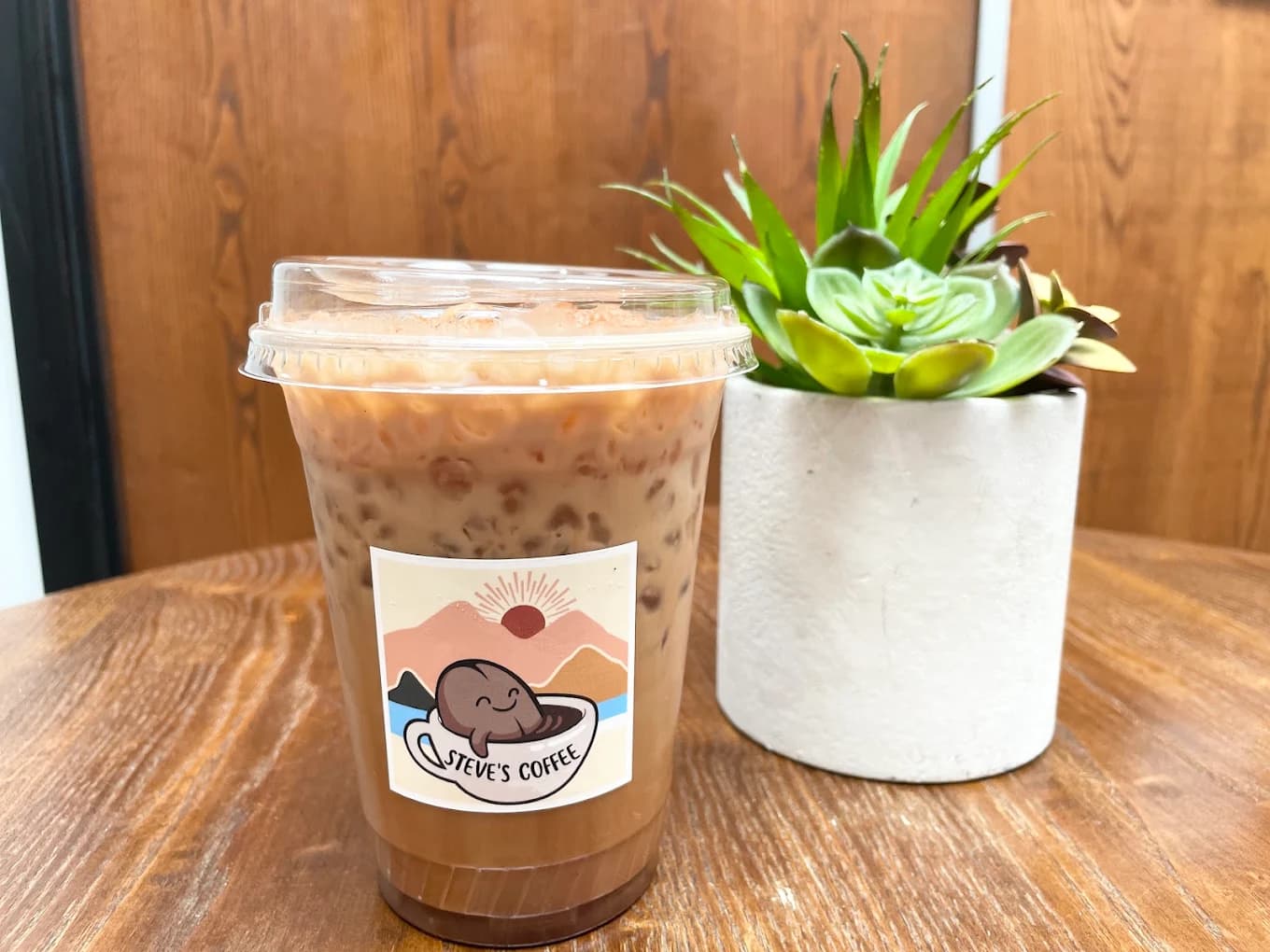 Iced coffee in a clear cup with the Steve's Coffee sticker beside a potted succulent on a wood table