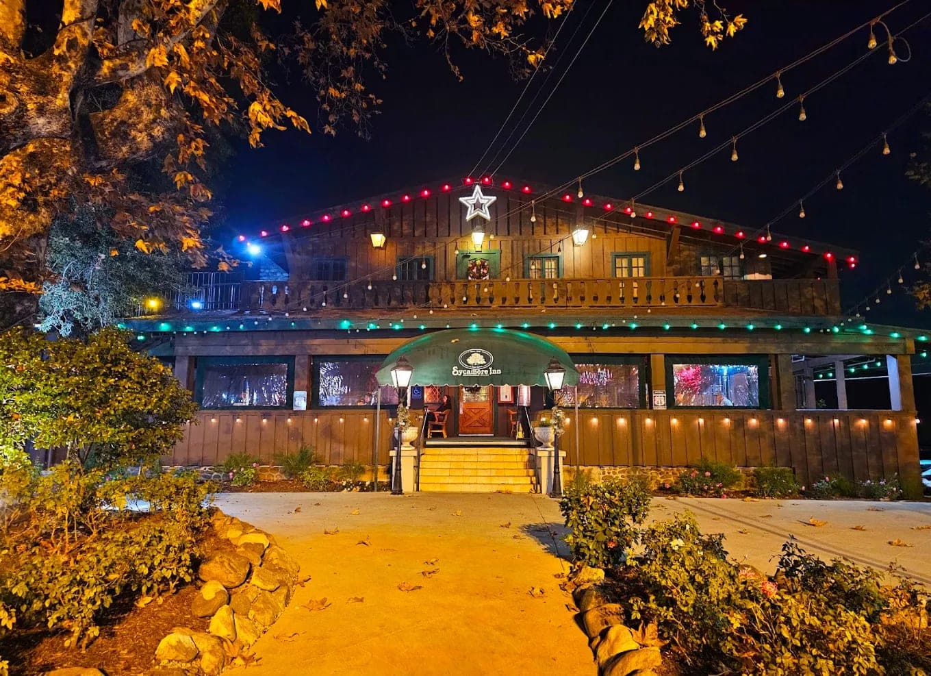 The Sycamore Inn at night, its timber lodge front strung with lights above a green awning over the entrance