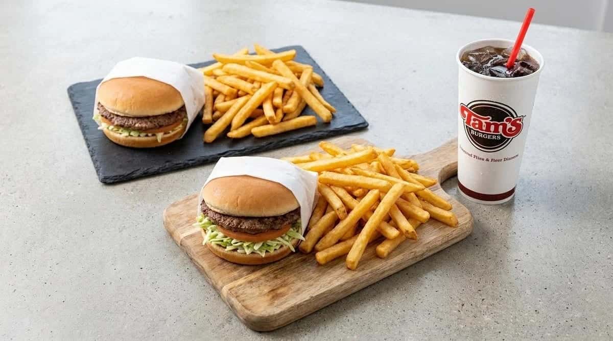 Two cheeseburgers with fries on a wooden board and a slate, beside a drink cup with a red straw