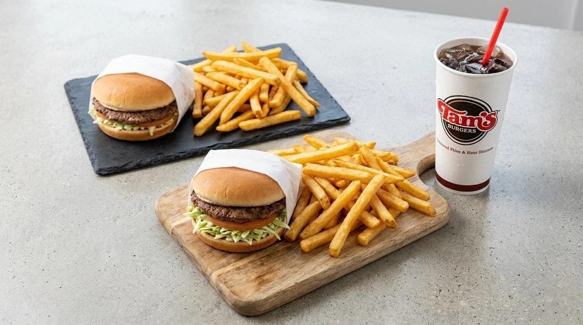 Two cheeseburgers with fries on a wooden board and a slate, beside a drink cup with a red straw