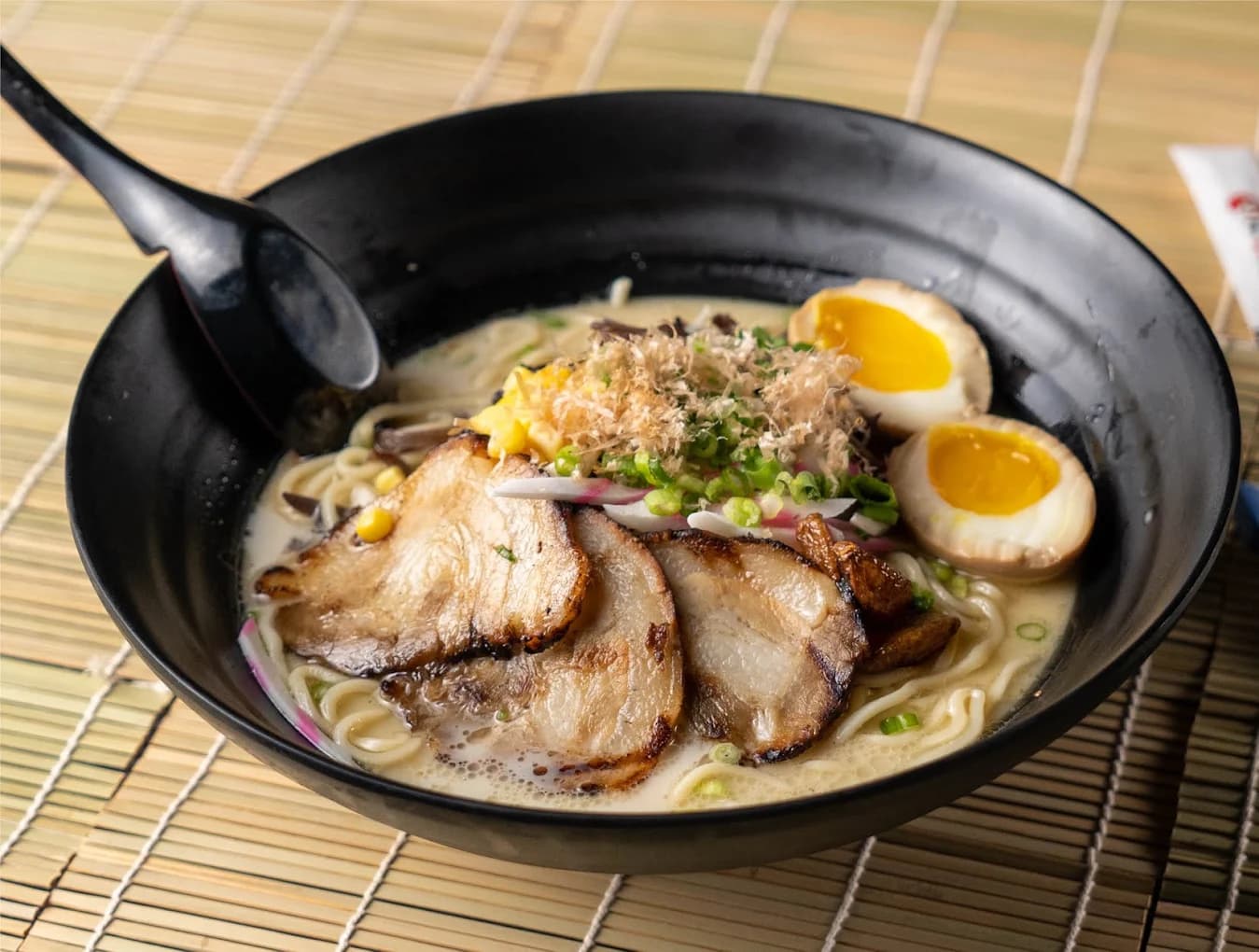 Black bowl of ramen with sliced pork, soft-boiled eggs and green onion on a bamboo mat at Tasty Pot