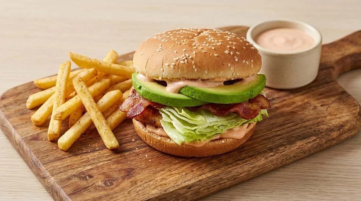 A bacon and avocado burger on a sesame bun with fries and a cup of pink sauce on a wooden board