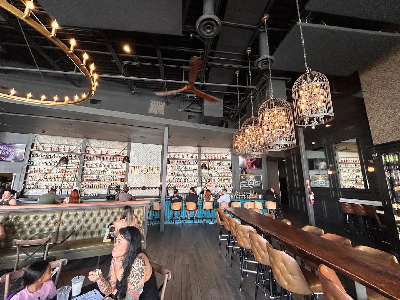 Bar room at The State with a back wall of liquor bottles, birdcage chandeliers and leather stools