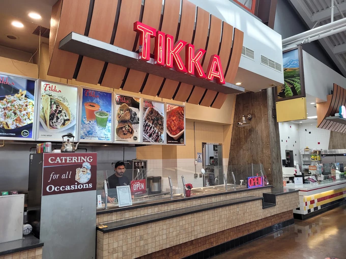Food court counter at Tikka with a red neon sign, photo menu boards and a catering sign