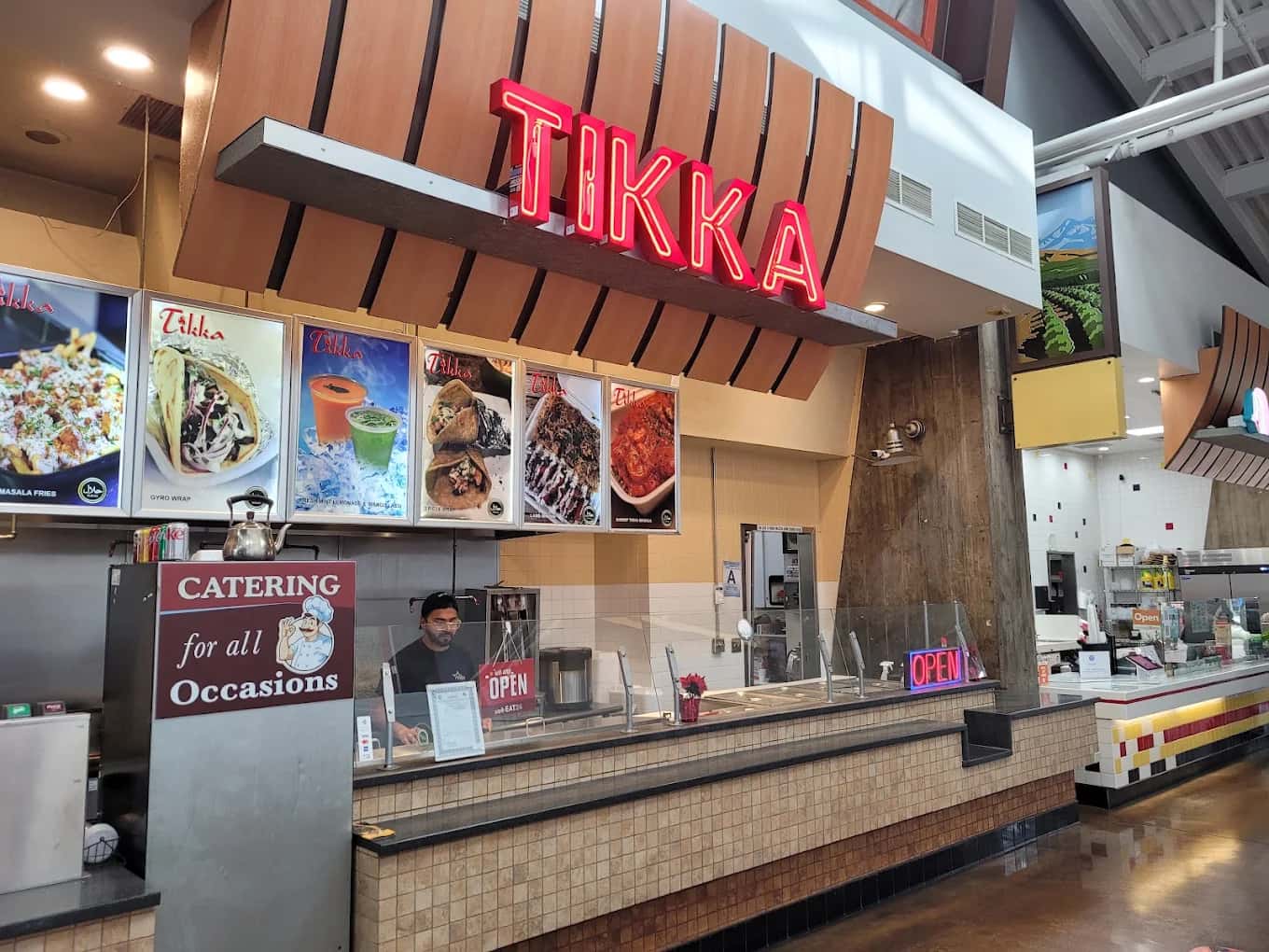 Food court counter at Tikka with a red neon sign, photo menu boards and a catering sign