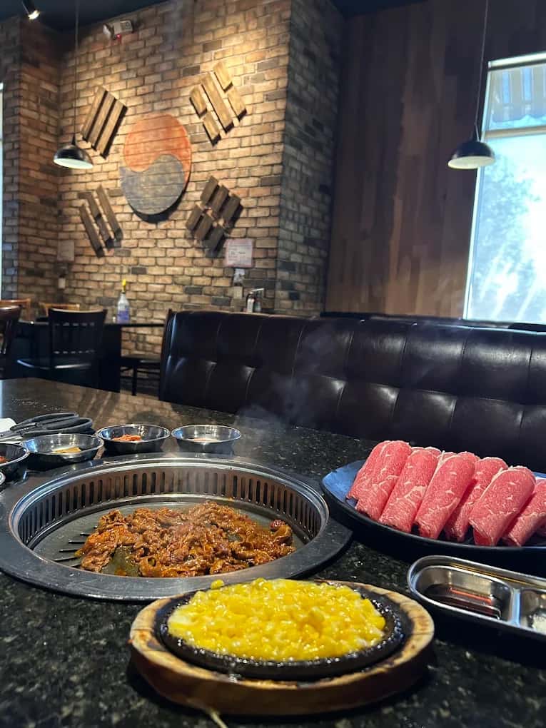 Tabletop grill at U Grill Korean BBQ with marinated meat, sliced beef, corn cheese and a brick wall behind