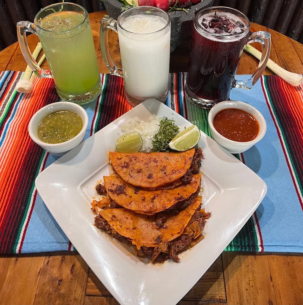 Plate of birria tacos with salsas and three mugs of aguas frescas at Victoria's Mexican Grill