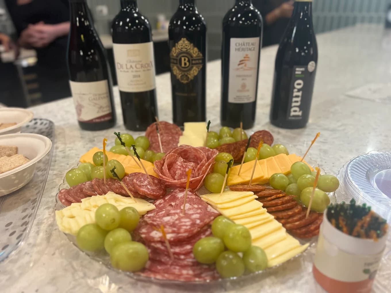 Platter of cured meats, cheese and grapes in front of five wine bottles on the counter at Wine Now!