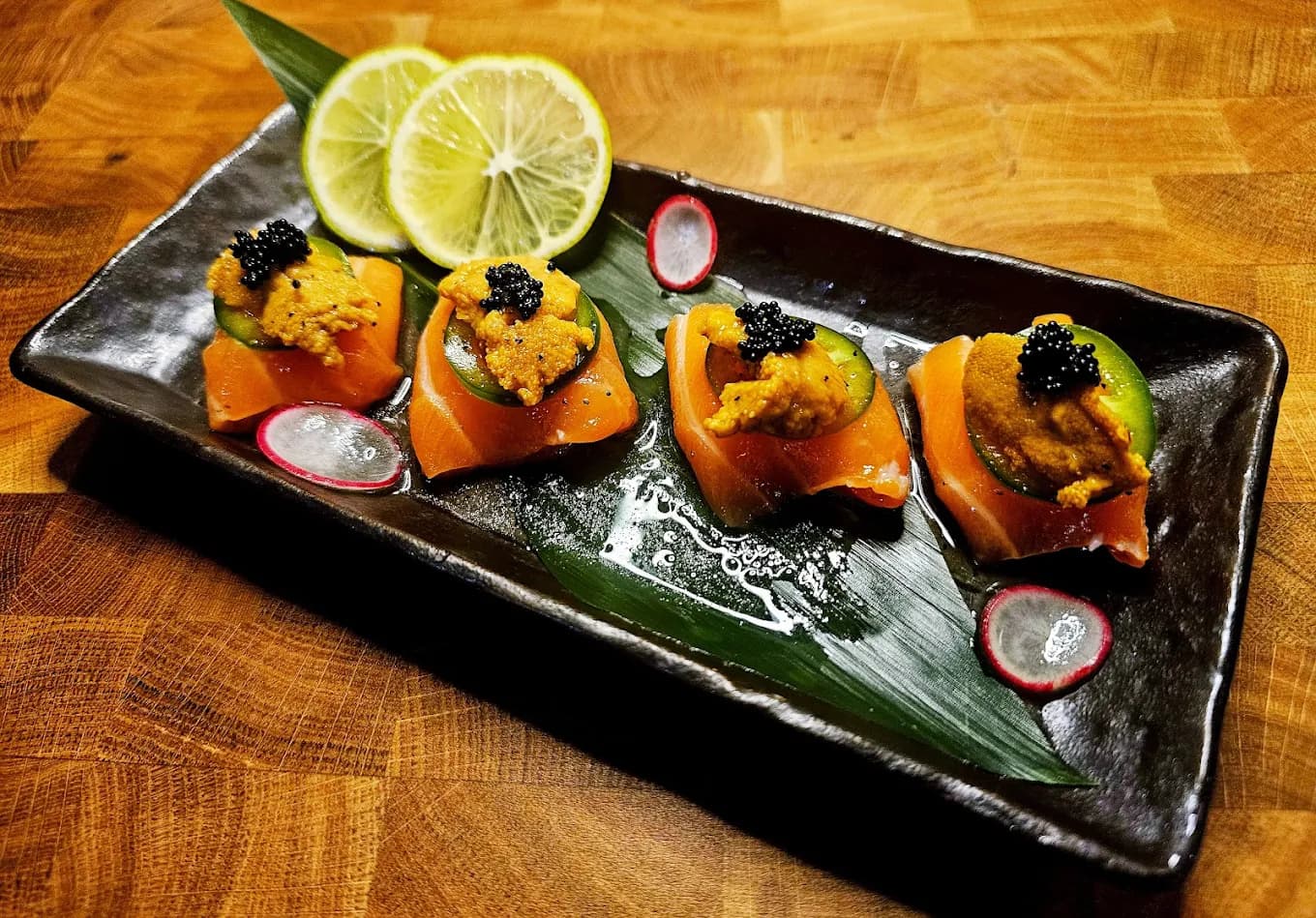 Four pieces of salmon topped with jalapeno, spicy sauce and black roe on a dark plate at Yatai Sushi