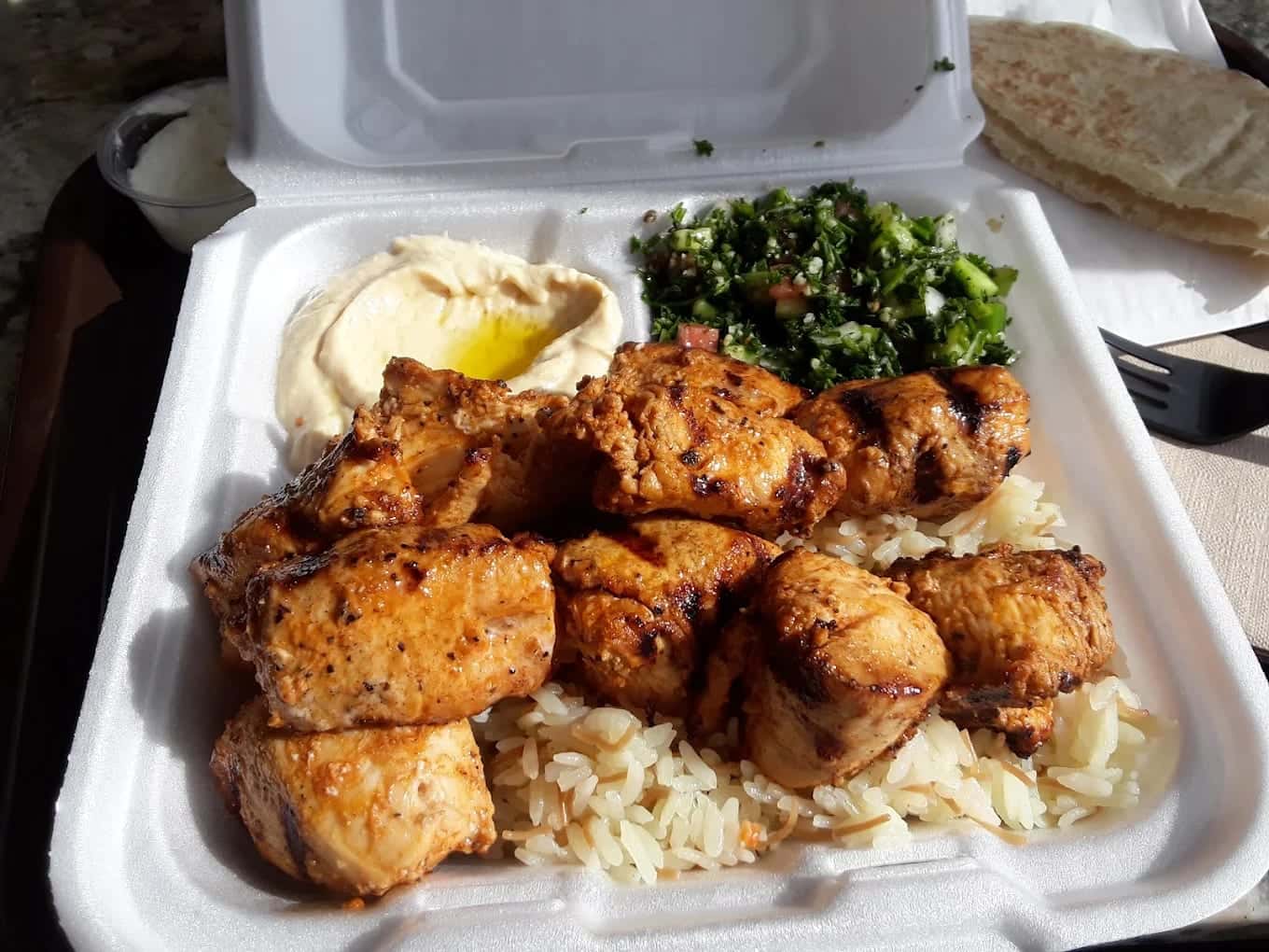 A takeout tray of grilled chicken over rice with hummus, tabbouleh and pita alongside