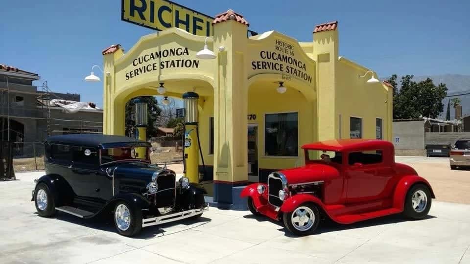 Two hot rods parked in front of the Cucamonga Service Station, a black Model A beside a red coupe under the Richfield sign